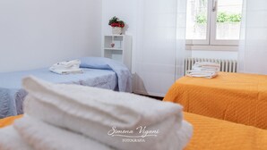 A hotel room with two beds, one with a blue bedspread and the other with an orange bedspread. There is a white radiator, a white shelf with a plant, and a window with a view of greenery outside. - bright and spacious apartment with large terrace overlooking the lake (Lovere)