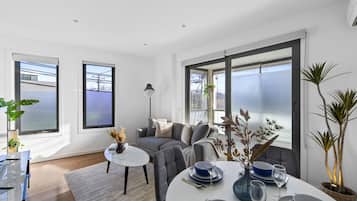 A modern living room with a grey sofa, a round dining table set with blue plates, and large windows with blinds.