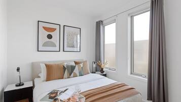 A hotel room with a bed, a nightstand, a window with curtains, and two framed artworks on the wall.