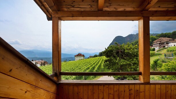 A wooden balcony with a view of a vineyard, a mountain, and a building in the distance.