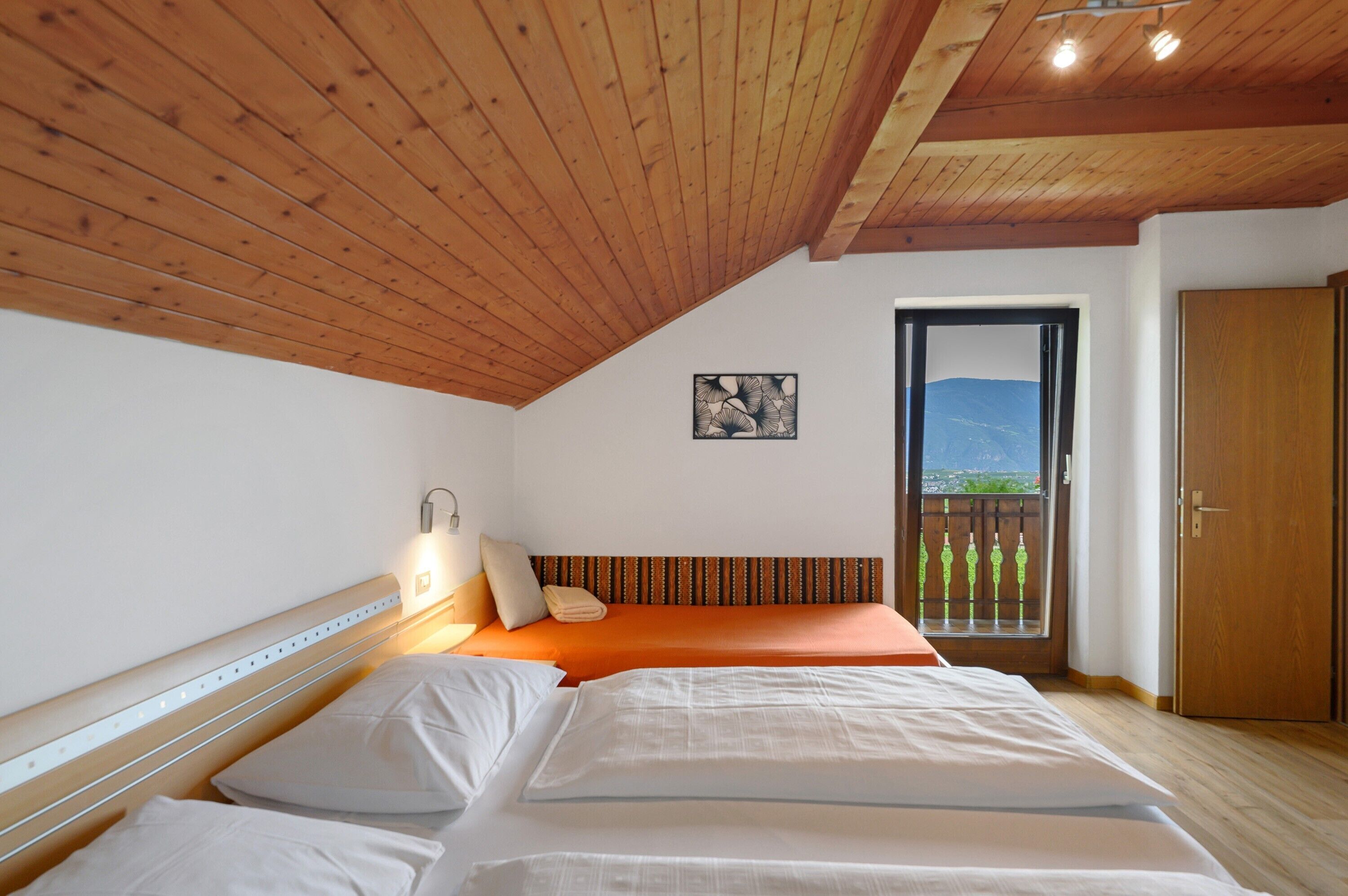Ein Schlafzimmer mit Holzbalkendecke, einem Bett mit weißer und orangefarbener Bettwäsche, einem Nachttisch mit Lampe, einem Fenster mit Bergblick und einer Tür.