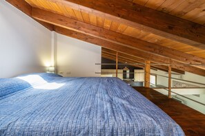 Une chambre avec un grand lit, des poutres apparentes en bois au plafond, une balustrade en verre et un escalier.