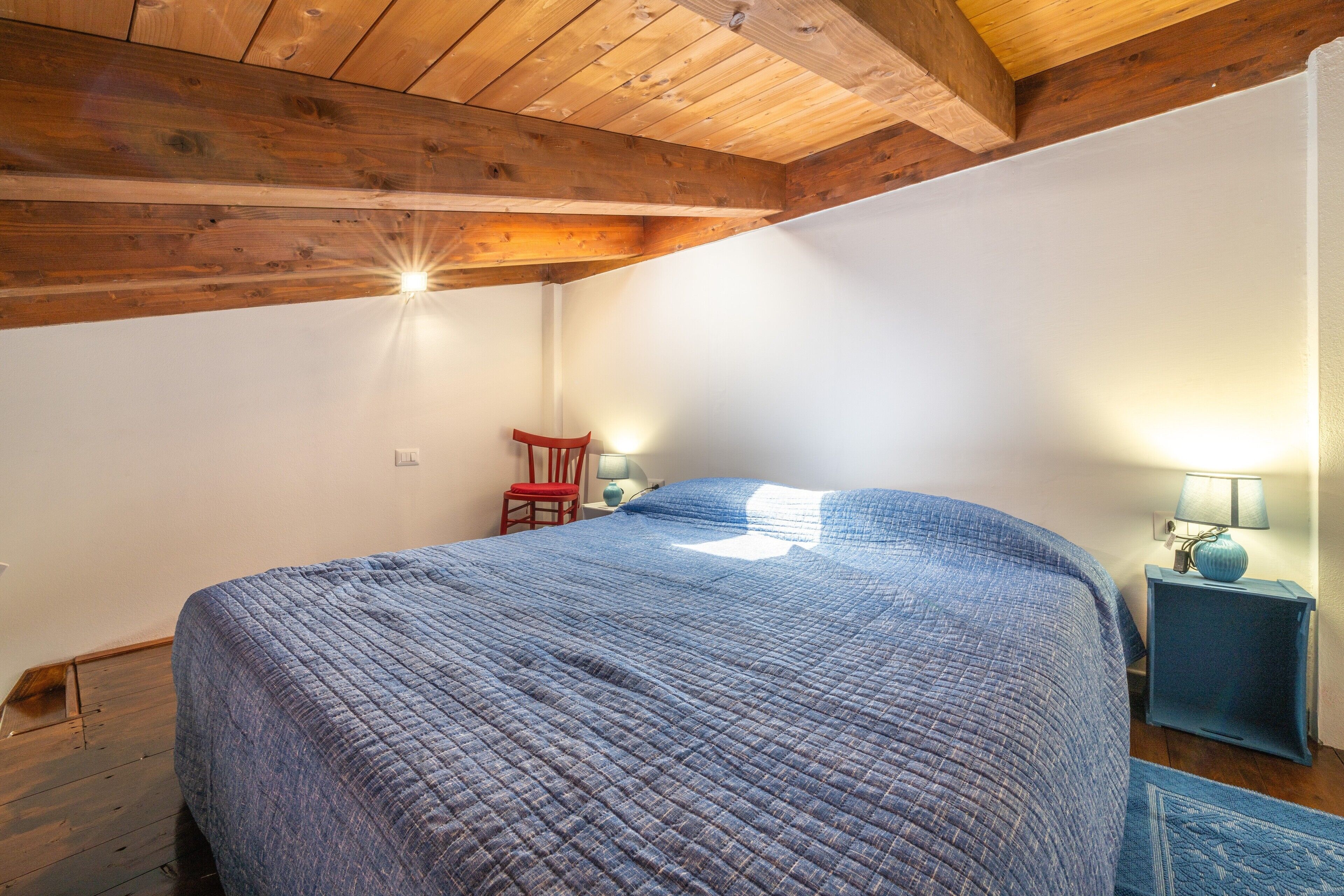 A bedroom with a wooden ceiling, a bed with a blue quilt, a red chair, and a nightstand with a lamp.