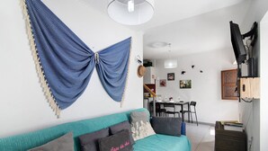 A living room with a sofa, a dining area, a TV, and a wall-mounted lamp. - Holiday Home 'Casa Da Avó Edite' with Private Terrace, Wi-Fi & Air Conditioning (Tavira)