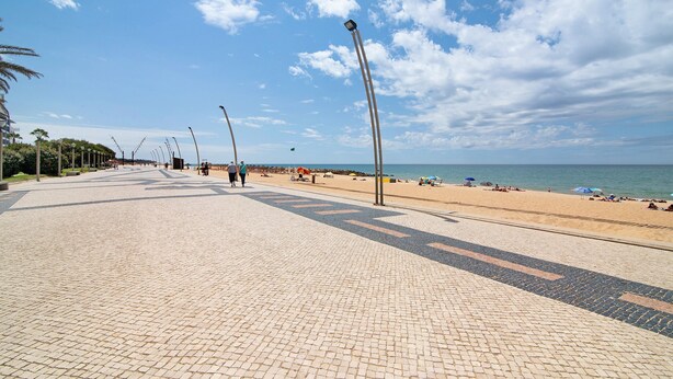 Ein gepflasterter Promenadenspaziergang führt zu einem Strand mit Sonnenschirmen und Menschen.