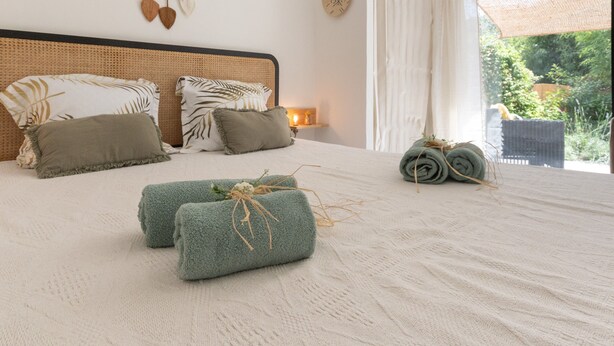 A bed with a woven headboard, two pillows, a bedside lamp, and rolled towels on the floor. - Holiday Gîte 'T1 Ardeche Vesseaux' with Mountain View, Shared Garden & Wi-Fi (Vesseaux)