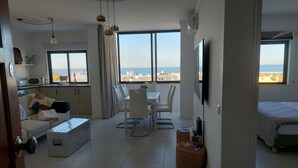 A modern living room with a dining area, large windows with a city view, a flat-screen TV, and a kitchenette.