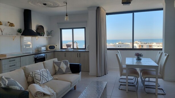 A modern living room with a sofa, dining table, chairs, and a view of the sea.
