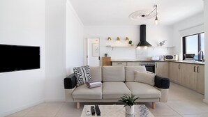 A modern living room with a sofa, coffee table, and TV. There is a kitchen in the background with cabinets and a stove.