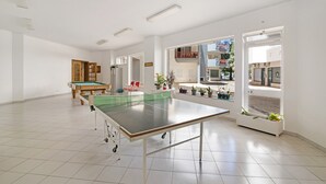 Een ruime kamer met een tafeltennistafel, een pooltafel en een deur naar een balkon met planten.