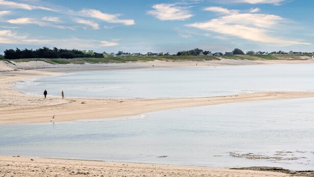 Una spiaggia con un’ampia zona sabbiosa e acqua calma, con alcune persone che passeggiano.
