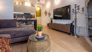 En moderne stue med en grå sofa, et rundt vævet sofabord med en potteplante, et fjernsyn monteret på væggen og et træspisebord med stole.
