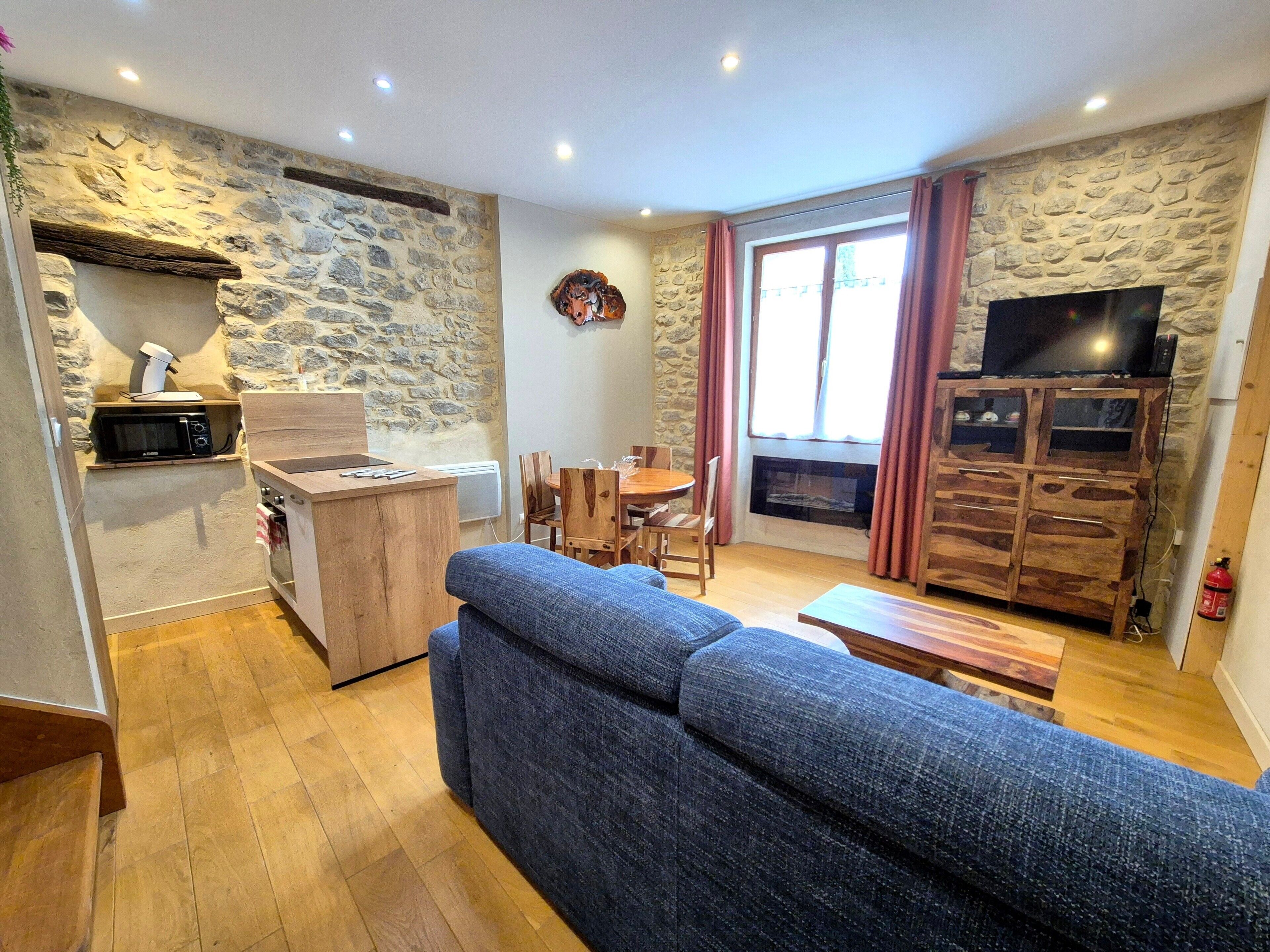 Un salon avec un mur en pierre, une table à manger en bois, un canapé bleu et un téléviseur posé sur un meuble en bois.