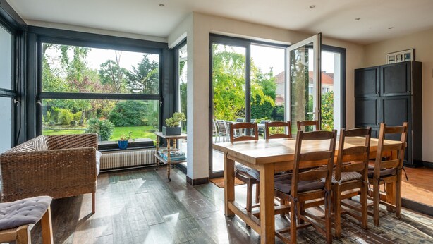 Un espace repas comprenant une table et des chaises, de grandes fenêtres et une vue sur la verdure.
