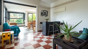 Une pièce avec un sol à carreaux, un canapé, une table, des chaises, une plante en pot et une vue sur l’extérieur par une porte ouverte.
