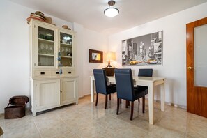 A dining area with a table, chairs, a cabinet with glass doors, a lamp, and a cityscape painting on the wall. - Apartment 'Atico Pepe Y Rosi' with Sea View, Wi-Fi and Air Conditioning (Cunit)