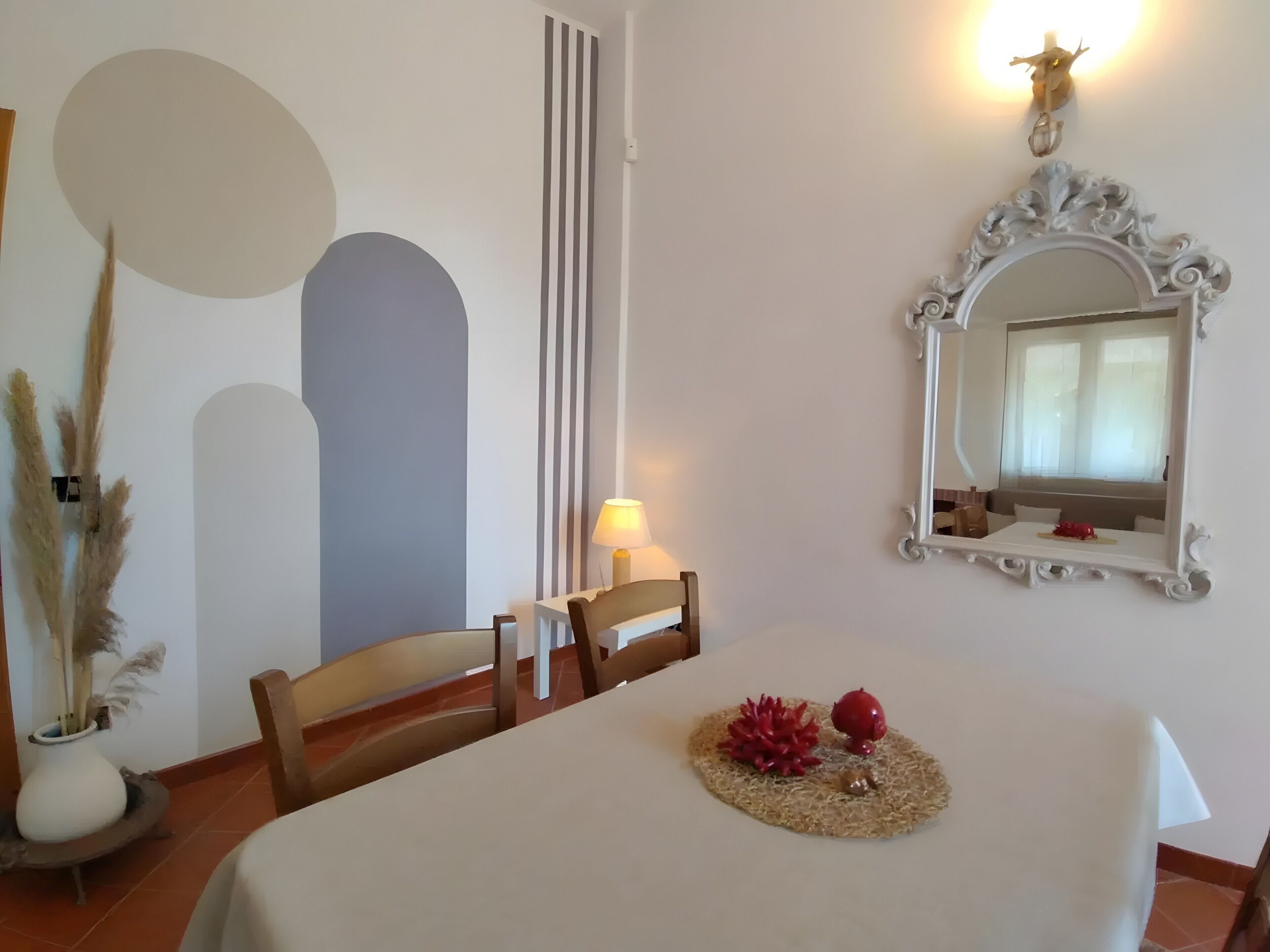 Un espace repas comprenant une table, des chaises, une lampe, des objets décoratifs, un miroir et un mur muni d’un luminaire.