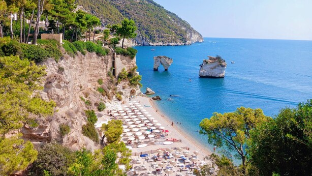A coastal beach with a rocky cliff, a paved walkway, and beach umbrellas. - Villa 'Meridiana Case Al Mare' with Sea View, Private Terrace and Air Conditioning (Mattinata)