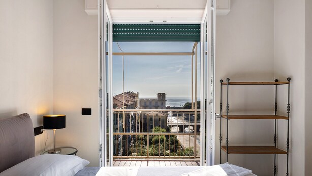 Ein Hotelzimmer mit Bett, Nachttisch, Lampe, einem Balkon mit Geländer und Blick auf Gebäude.