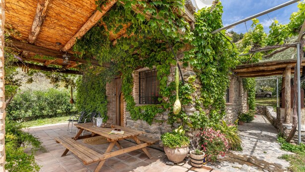 A rustic outdoor seating area with a wooden table and chairs, surrounded by potted plants and a stone pathway.