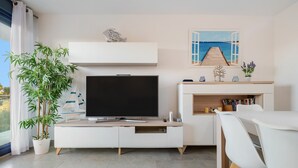 En moderne stue med et fladskærms-TV, et spisebord, hvide møbler, en stor stueplante, et maleri af en havn og udsigt gennem et vindue.