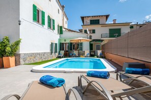 Une piscine entourée de bâtiments aux volets verts, avec des chaises longues et des parasols.