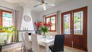 Comedor con una mesa blanca, cuatro sillas, un florero con flores, un ventilador de techo y puertas de madera con barrotes.