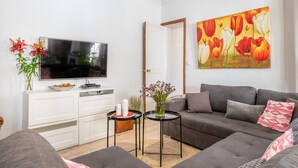 Una sala de estar con un televisor de pantalla plana, un sofá gris, un mueble blanco y un cuadro con flores en la pared.