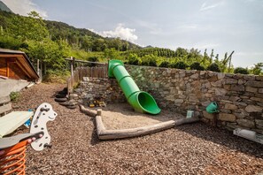 Une aire de jeux avec un toboggan vert, une balançoire en bois en forme de chien et un mur en pierre.