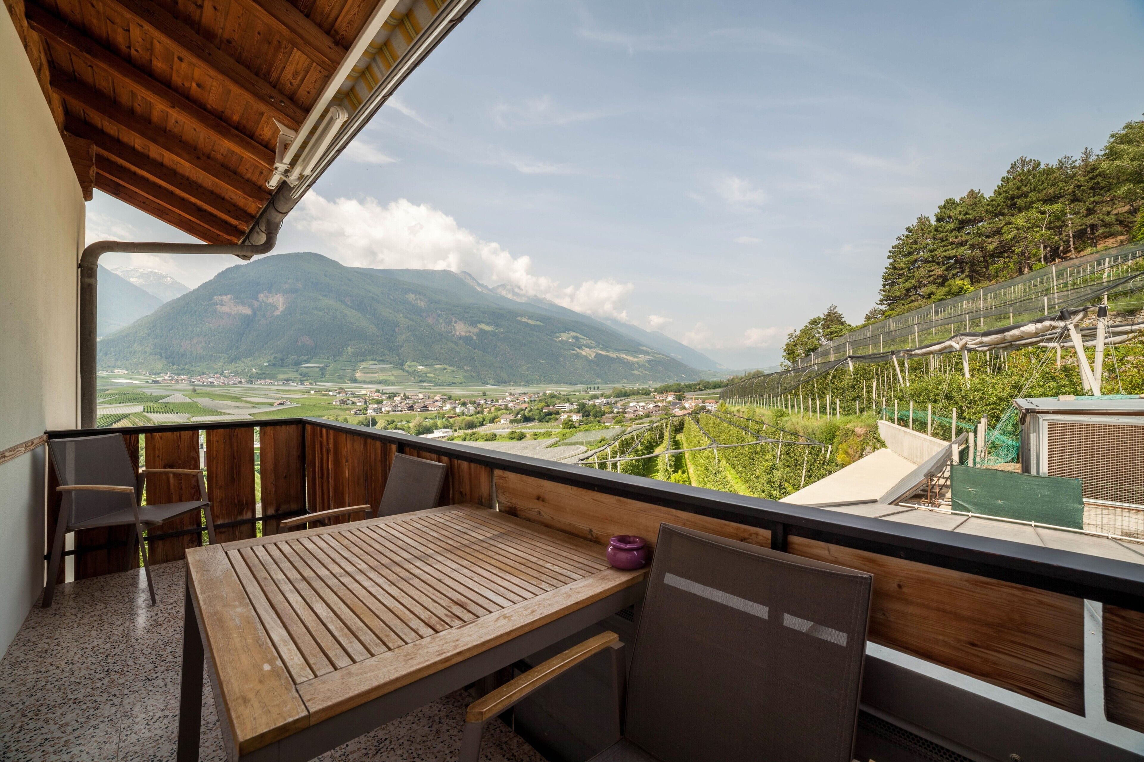 Ein Balkon mit Holztisch und -stühlen, Blick auf eine Stadt und Berge sowie ein grüner Zaun.