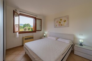 A bedroom with a bed, a nightstand, a wall-mounted lamp, a window with a view of greenery, and a framed picture on the wall. - Apartment 'Casa Vacanze Susino' with Shared Pool, Private Terrace and Air Conditioning (Olbia)