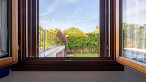 A window with a view of a balcony, a railing, and a garden with trees and flowers.