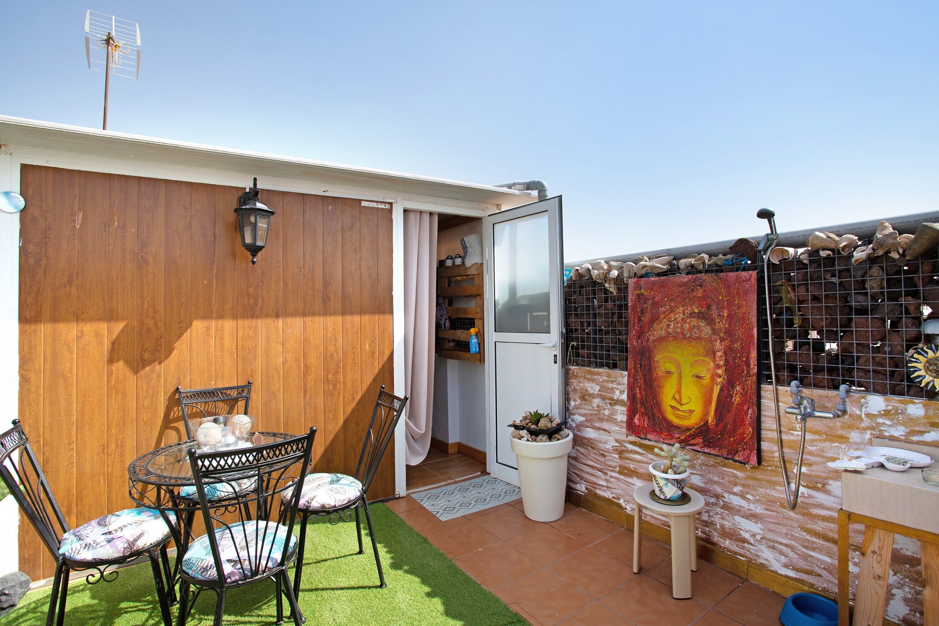 Un patio con una mesa y sillas, un taburete pequeño, una planta en maceta, una luz fijada en la pared y una puerta que da a un espacio interior.