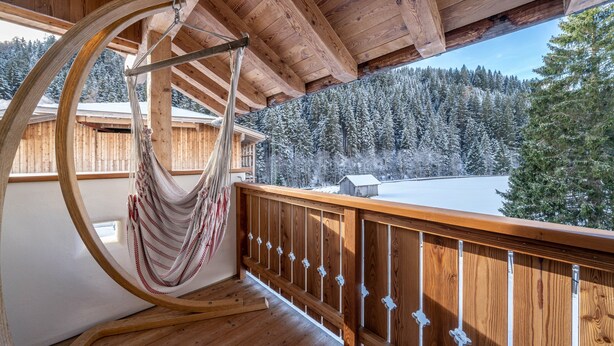 A wooden deck with a hammock, wooden railings, and a view of a snowy forest.
