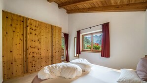 A bedroom with a wooden wardrobe, a bed with white bedding, a window with red curtains, and a view of mountains. - Holiday Apartment "Larch Soge Apt Helmuth" with Mountain View, Balcony & Shared Garden (Racines)