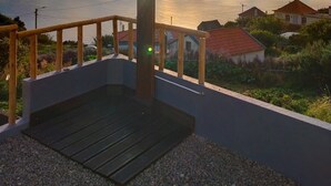 Een houten terras met een metalen hekwerk, een betonnen plateau en uitzicht op zee en huizen op de achtergrond.