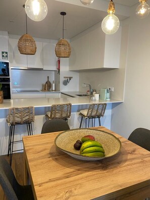 Une cuisine moderne avec des meubles blancs, un coin repas avec des chaises en osier et un panier de fruits sur la table.