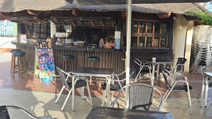 An outdoor café with metal tables and chairs, a thatched roof, and a bar area with a counter and stools.