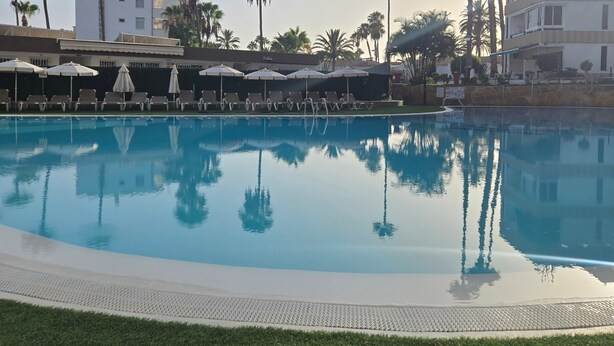 A swimming pool with lounge chairs and umbrellas, surrounded by palm trees and buildings.