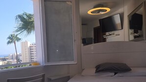 A hotel room with a bed, a chair, a window with a view of palm trees and buildings, a TV mounted on the wall, and a ceiling light fixture.