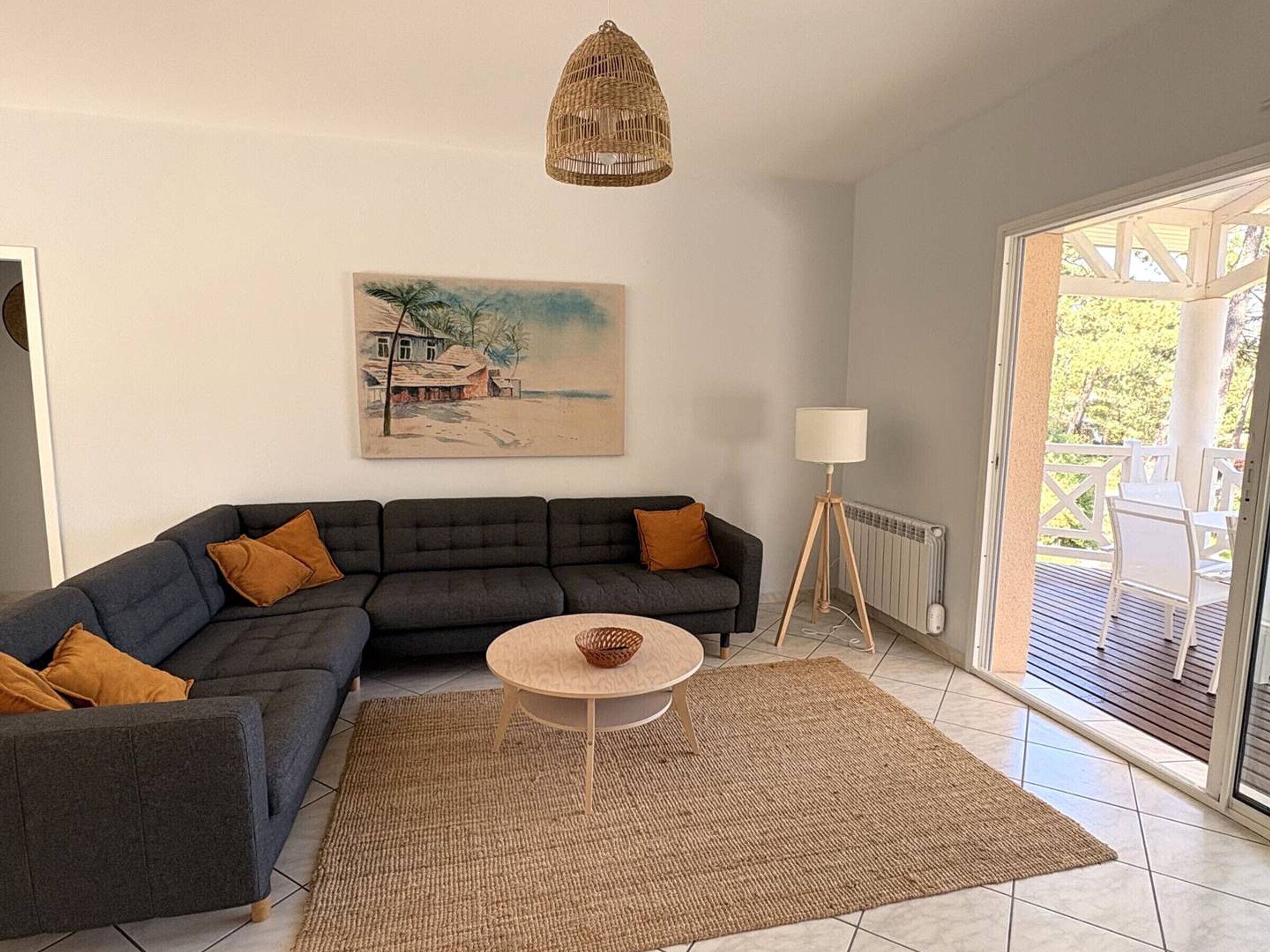 Ett vardagsrum med en mörk soffa, ett runt soffbord, en vävd matta, en tavla som föreställer en strandscen och en utsikt genom en glasdörr ut till en uteplats.