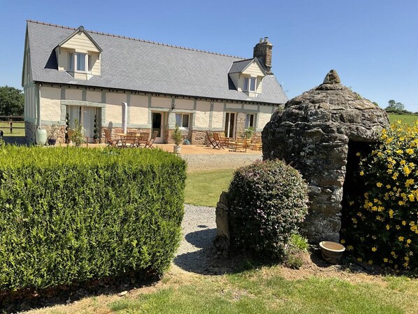 A stone house with a grey roof, a stone chimney, and a well-maintained garden with hedges and flowering plants.