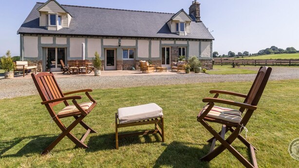 A two-story house with a stone facade, a large patio with wooden furniture, and a view of a rural landscape.