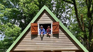 Une cabane en bois avec un toit triangulaire, une porte d’entrée et un petit porche équipé de chaises en plastique.