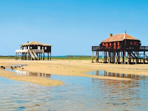 Dos estructuras de madera con balcones sobre pilotes, frente a una playa arenosa y agua.