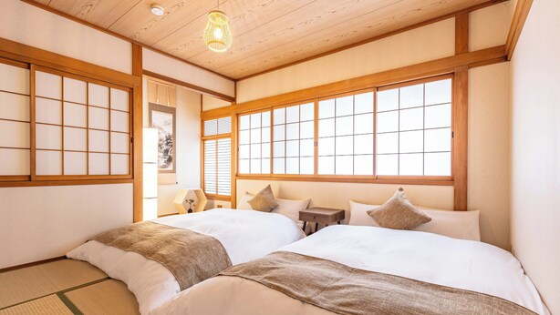 A traditional Japanese-style room with tatami flooring, two beds with white bedding, wooden sliding doors, and large windows with shoji screens. - Precious House Nipponbashi (Osaka)