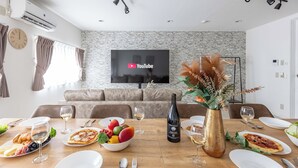 A modern dining area with a wooden table set for a meal, a television mounted on the wall, and a stone-textured wall behind the seating. - Precious House Nipponbashi (Osaka)