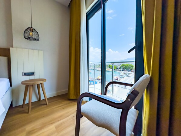 A hotel room with a bed, a wooden chair, a small round table, and a large window with a view of a marina and boats.