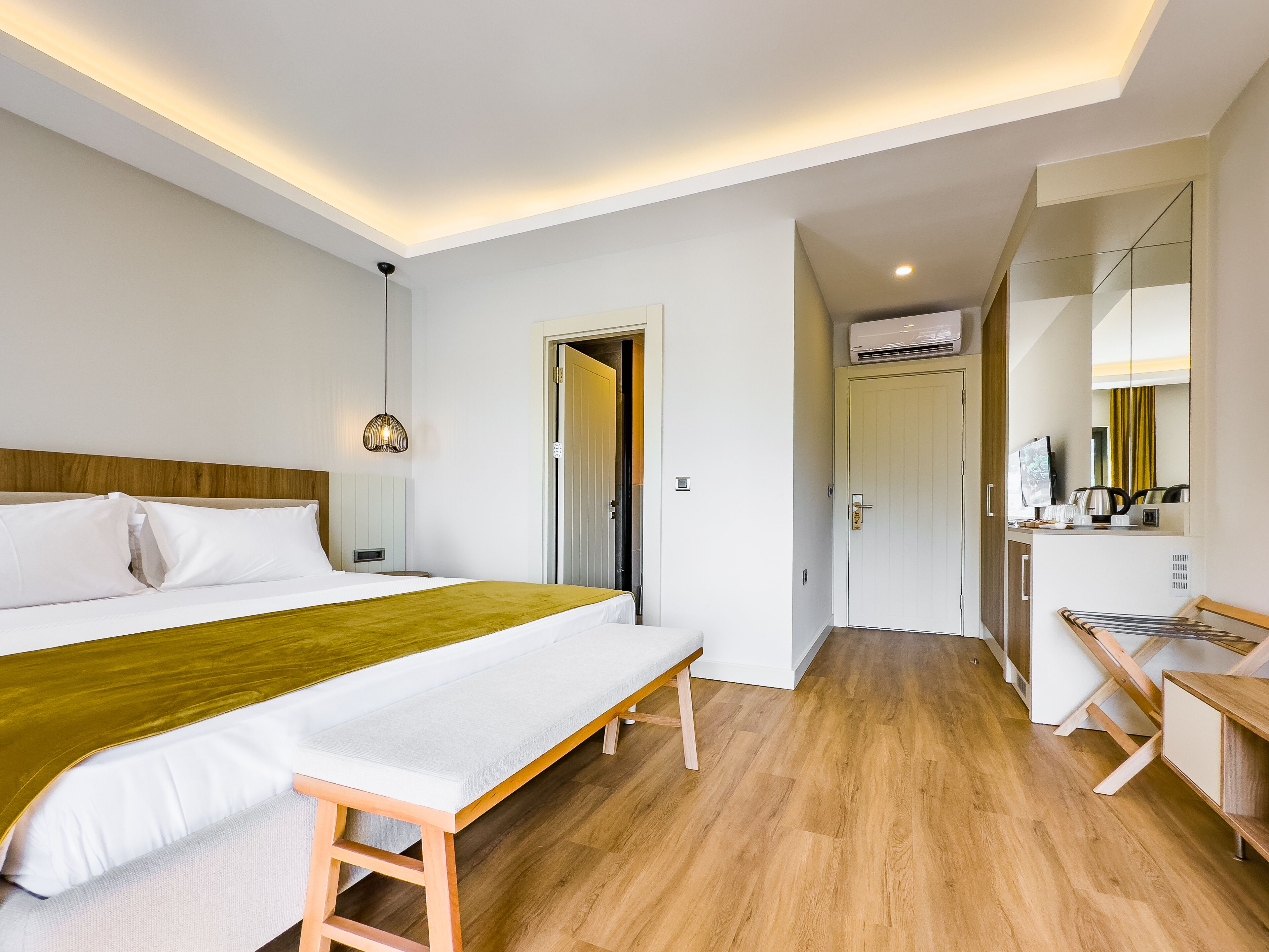 A modern hotel room with a large bed, wooden flooring, a bench, a desk, and a mirror.