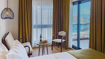 Habitación de hotel con una cama, dos sillas, una mesita con una botella y vasos, y vistas a un puerto deportivo a través de amplias ventanas.